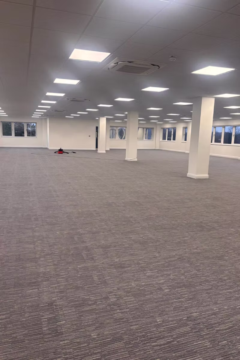 Large empty office room with gray carpet tiles, white pillars, and square ceiling lights.