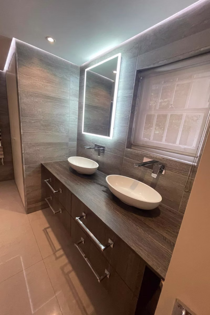 Bathroom vanity with two white vessel sinks, gray tiled walls, and an illuminated mirror.