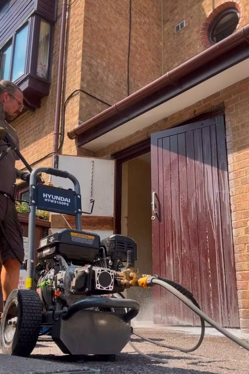 High-pressure washing machine spraying water to clean a wooden exterior door of a brick building.